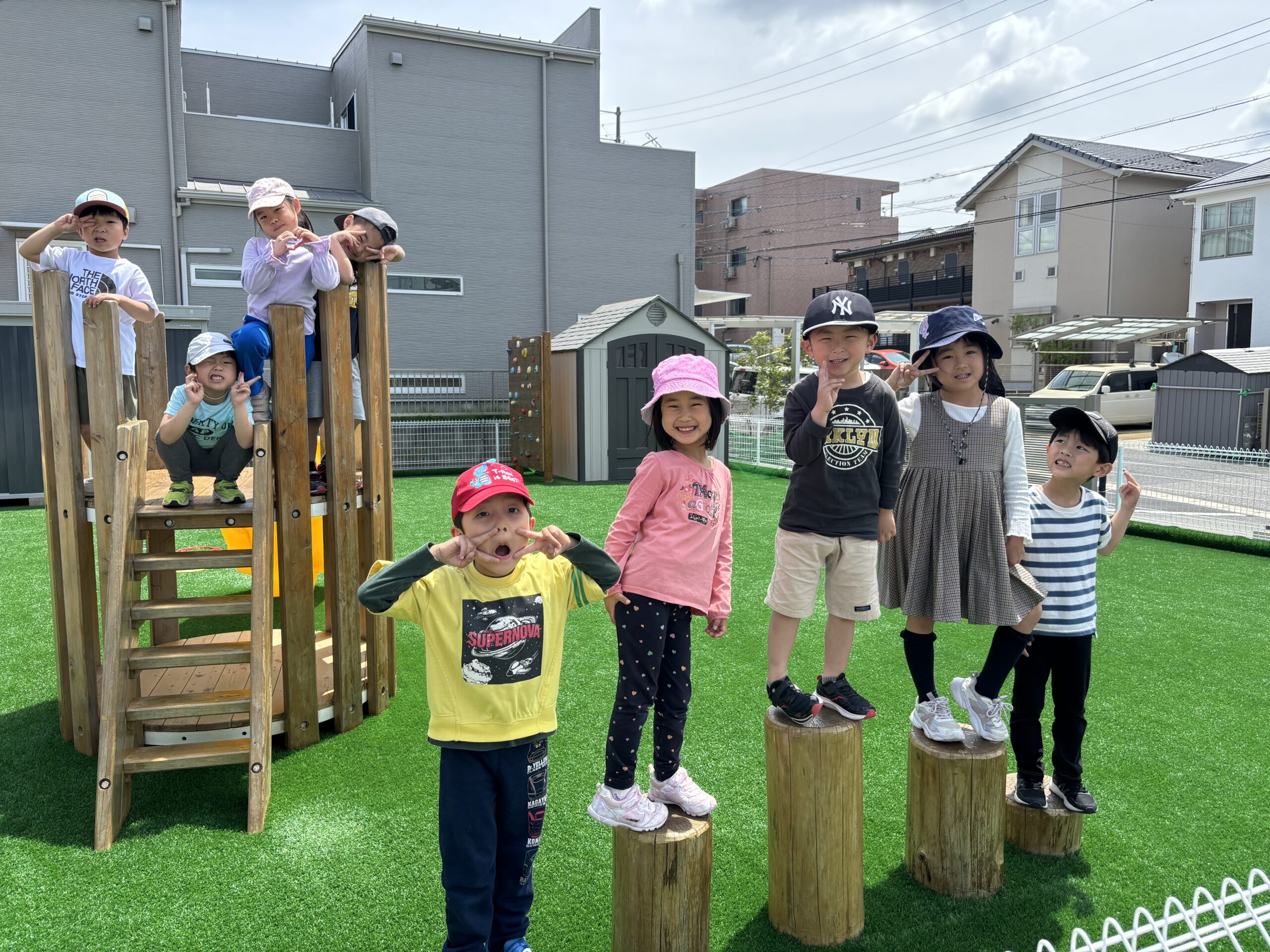 園庭でポーズをとる子どもたち|ディスカバリーインターナショナルスクール 名古屋のディスカバリーインターナショナルスクールの園庭で、楽しそうにポーズをとる幼稚園児たち。木製の遊具と広い屋外スペースが、安全な国際スクールの環境で身体の発達を促します。