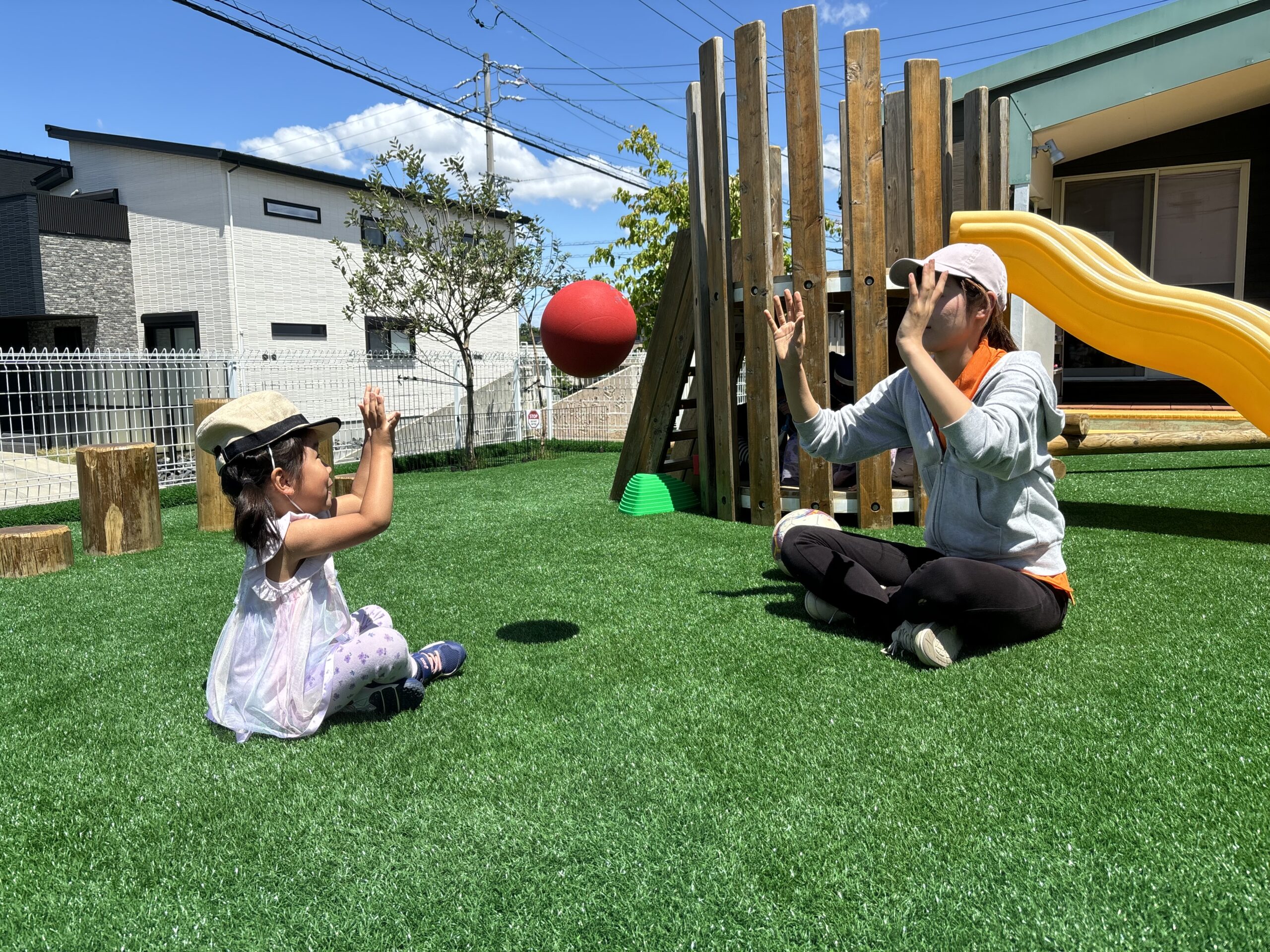 長久手の屋外遊び場で先生とボール遊びをする子ども|ディスカバリーインターナショナルスクール ディスカバリーインターナショナルスクール(名古屋・長久手)の園庭で、先生と赤いボールを使ってキャッチボールをする幼児の様子。プレイベースの学びを支える屋外施設の一部。